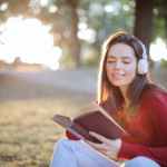 selective focus photo of smiling woman in a red long sleeve top reading book while listening to music on headphones