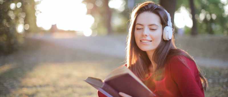selective focus photo of smiling woman in a red long sleeve top reading book while listening to music on headphones