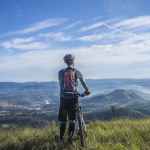 biker holding mountain bike on top of mountain with green grass